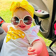 Kali is registered to the contest to win money with this photo: baby, child, sunglasses, yellow_headwrap, white_shirt, car_door, grass, smiling, happy, outdoor, casual, infant, sunlight, portrait, cute, fashion_accessory, seat, hand, candid, summer