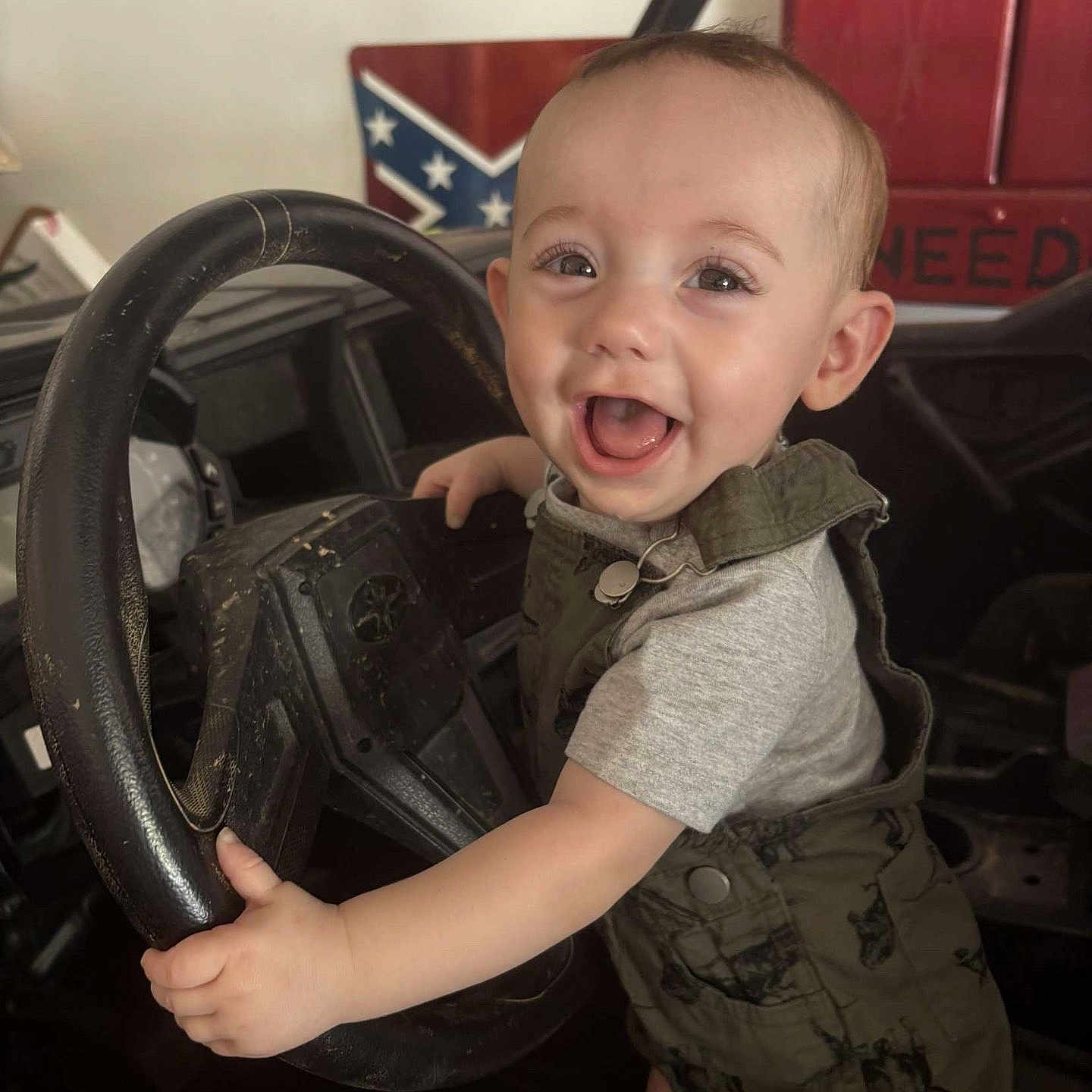 Dawson is registered to the contest to win money with this photo: baby, cabinet, child, clothing, eyes, face, flag, garage, hand, happy, indoor, joyful, overalls, person, playful, portrait, rustic, smile, steering_wheel, vehicle