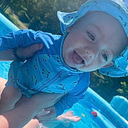 Dawson is registered to the contest to win money with this photo: baby, child, blue_clothing, sun_hat, pool, water, inflatable_pool, outdoor, summer, playful, tongue_out, person, sunny, trees, sky, greenery, happy, hands, swimsuit, toys