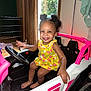 Amaya is registered to the contest to win money with this photo: toddler, child, toy_car, indoor, smiling, barefoot, yellow_dress, pink, white, black_hair, happy, steering_wheel, window, curtains, floor, door, play, fun, person, furniture
