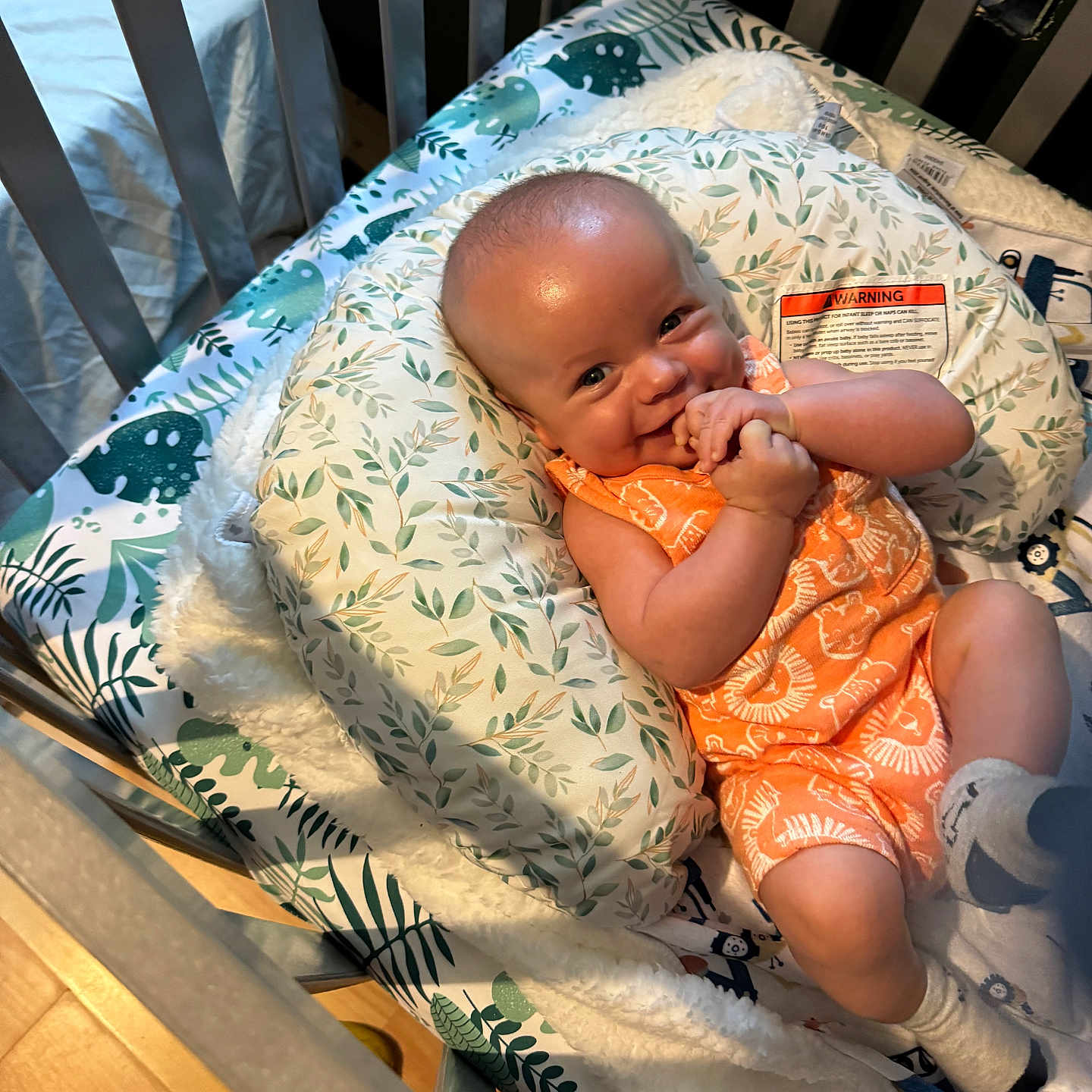 Grayson is registered to the contest to win money with this photo: baby, baby_bedding, baby_nest, blanket, child, comfort, crib, cute, hand_clasp, happy, indoor, infant, leaf_pattern, orange_clothing, person, playful, romper, smile, socks, wooden_floor