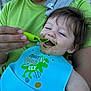 Dante is registered to the contest to win money with this photo: baby, child, feeding, bib, green_food, messy, smile, happy, adult, hand, spoon, clothing, indoor, person, face, hair, blue_bib, green_shirt, closeup, mealtime
