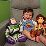 toddler, child, smile, toy_story, buzz_lightyear, woody, plush_toys, armchair, blue_overalls, happy, indoor, green_wall, window, light, portrait, cute, young_child, sitting, clothing, foot