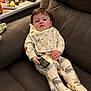 Dante is registered to the contest to win money with this photo: baby, child, remote_control, pajamas, couch, toy, giraffe_plush, table, blanket, window_blind, clothing, face, hand, furniture, indoor, person, sitting, expressionless, home, infant