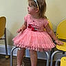 child, toddler, girl, portrait, tiara, pink_dress, tutu, sitting, chairs, classroom, indoor, smiling, profile, blonde_hair, ponytail, shoes, wood_floor, wall, furniture, playroom