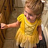 child, toddler, girl, portrait, smiling, happy, yellow_dress, hair_bow, blonde_hair, barefoot, indoor, kitchen, wooden_cabinets, tile_floor, rug, standing, looking_up, dress, bow, family_photo