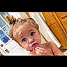 child, toddler, girl, portrait, face, smiling, eating, cake, frosting, dessert, messy_face, food, indoors, door, wooden_cabinet, bun_hair, hand, eyes, bare_shoulders, black_border