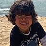 child, smiling, beach, sand, water, ocean, sunlight, waves, curly_hair, casual_clothing, tshirt, outdoor, happy, portrait, person, daylight, nature, fun, vacation, play