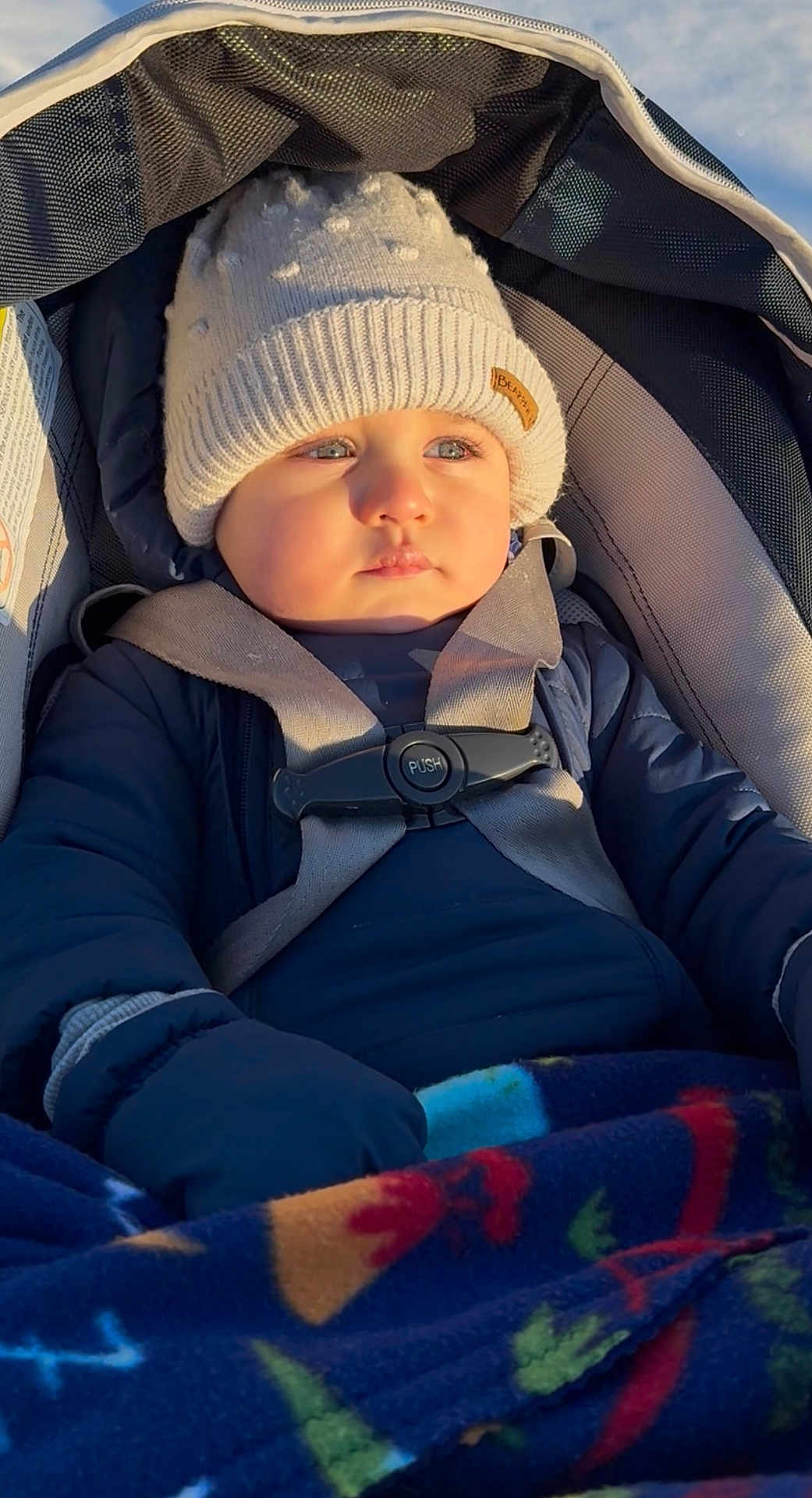 Axel is registered to the contest to win money with this photo: baby, child, car_seat, hat, winter_clothing, blanket, gloves, jacket, blue_eyes, outdoor, sunlight, warm, infant, portrait, seat_belt, cute, young, face, person, bundle