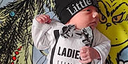Taylon is registered to the contest to win money with this photo: baby, infant, child, clothing, hat, blanket, grinch, cartoon, face, person, cute, smiling, white_shirt, patterned_pants, lying_down, indoors, small_hands, expression, colorful, funny_outfit