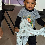 Djahnaël a rejoint le concours — aidez-le/la à gagner de superbes lots ! toddler, child, indoor, blanket, braided_hair, shirt, black_pants, floor, curious, standing, person, home, furniture, table, wall, cable, expression, young_child, clothing, portrait