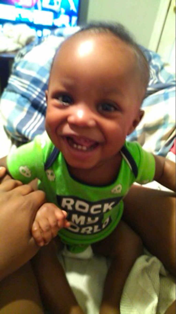 Rayjarious joined the competition — help win amazing prizes! bed, blanket, child, clothing, comfort, cute, face, green_shirt, hand, happy, indoor, infant, joyful, person, plaid, room, sitting, smiling, toddler, young_child