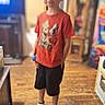 child, boy, indoor, standing, red_tshirt, black_shorts, white_sneakers, wooden_floor, blurred_background, furniture, shelf, casual_pose, short_hair, smiling, home, floor, person, clothing, toy, colorful_background