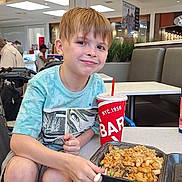 Brayden joined the competition — help win amazing prizes! child, boy, smile, food, fried_rice, shrimp, takeout_container, table, dining_area, mall, food_court, casual_clothing, sneakers, indoor, people, background, light, drink_cup, red_straw, seated