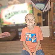 Desmond is registered to the contest to win money with this photo: child, boy, smile, orange_shirt, jeans, indoor, seated, casual, furniture, television, blurred_background, wood_floor, home, happy, portrait, person, young, short_hair, under_armour, confident