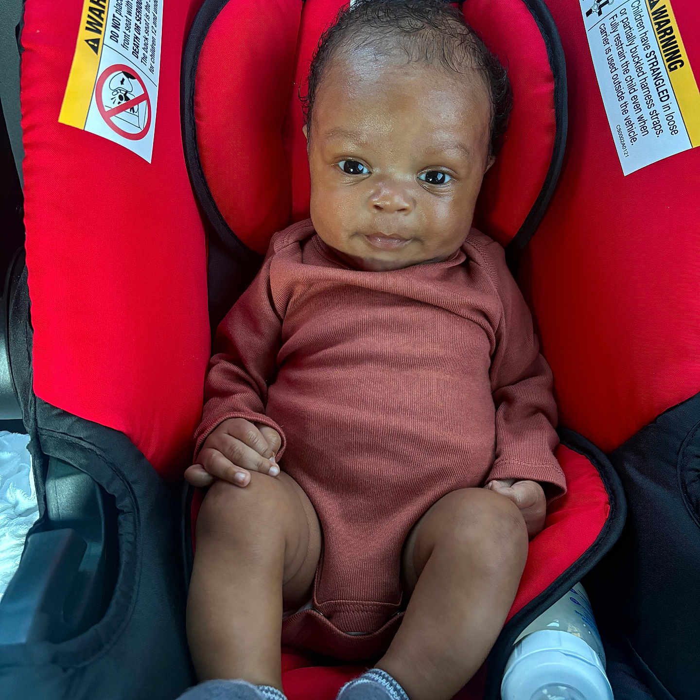 Nyaire is registered to the contest to win money with this photo: baby, infant, car_seat, red, onesie, socks, bottle, cute, child, portrait, indoors, seat, small, person, newborn, clothing, baby_bottle, face, looking, resting