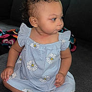 Laniyah is registered to the contest to win money with this photo: toddler, child, curly_hair, blue_dress, daisy_pattern, sitting, indoor, couch, gray_couch, curious_expression, barefoot, cross_legged, person, home_interior, blanket, curtains, natural_light, portrait, young_child, cute