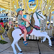 Nathael a rejoint le concours — aidez-le/la à gagner de superbes lots ! child, carousel, horse, amusement_ride, blue_cap, shorts, sandals, smiling, outdoor, daytime, fairground, wooden_floor, colorful, people, toy, fun, summer, playful, ride, happy