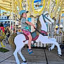 child, carousel, horse, amusement_ride, blue_cap, shorts, sandals, smiling, outdoor, daytime, fairground, wooden_floor, colorful, people, toy, fun, summer, playful, ride, happy