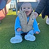 baby, child, smiling, sitting, playground, slide, vest, blue_sweater, white_shoes, grass, outdoor, daylight, happy, person, supporting_hand, climbing_structure, casual_clothing, fun, cute, young