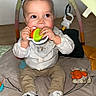 baby, child, toy, play_mat, play_gym, indoor, sitting, cute, messy_face, white_shirt, beige_pants, striped_shoes, floor, wooden_floor, colorful_toys, infant, hands, face, chewing, curious