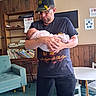 Aerial joined the competition — help win amazing prizes! man, baby, indoor, bookshelf, books, armchair, table, chair, carpet, cap, glasses, wood_paneling, holding, swaddled, person, infant, casual_clothing, reading_material, furniture, warmth