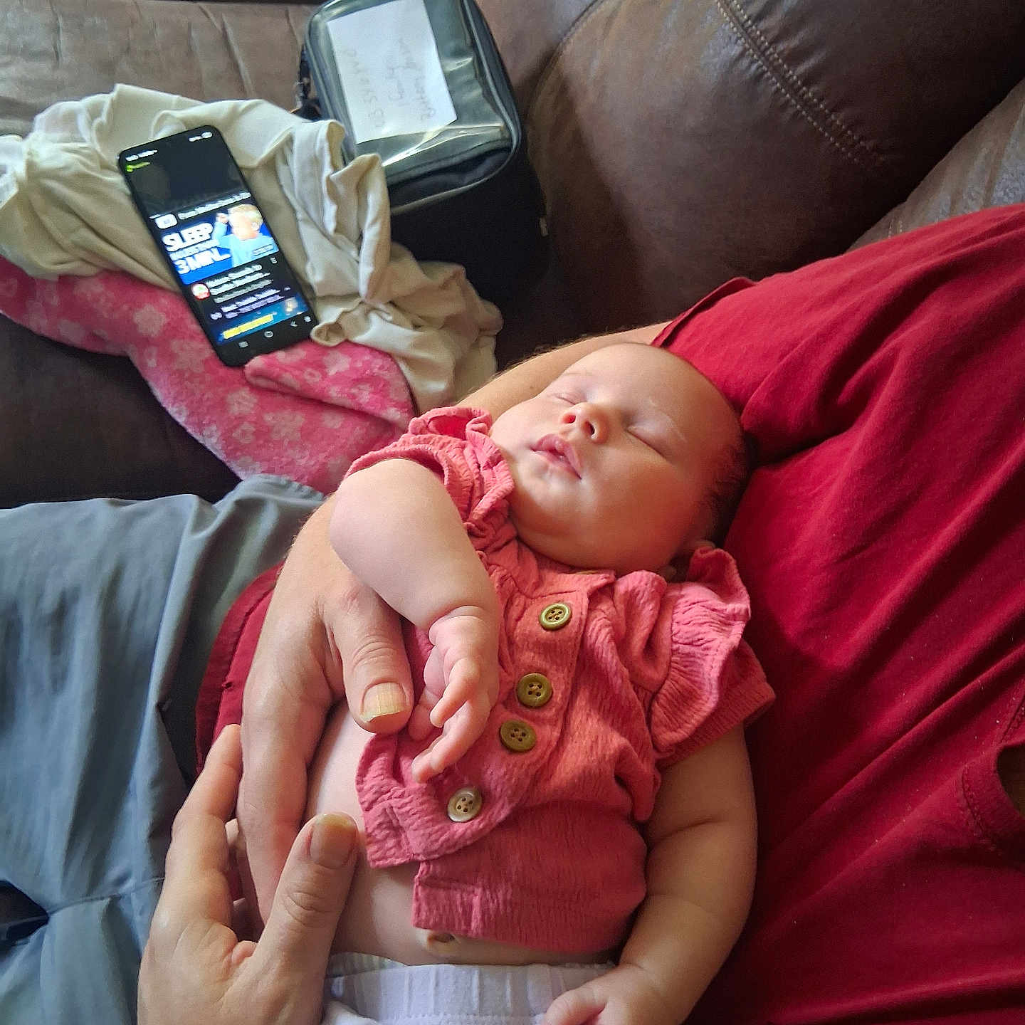 Aerial is registered to the contest to win money with this photo: baby, sleeping, infant, person, hand, clothing, red_shirt, pink_outfit, couch, sofa, phone, video, blanket, arm, fingers, relaxation, indoor, comfort, casual, home
