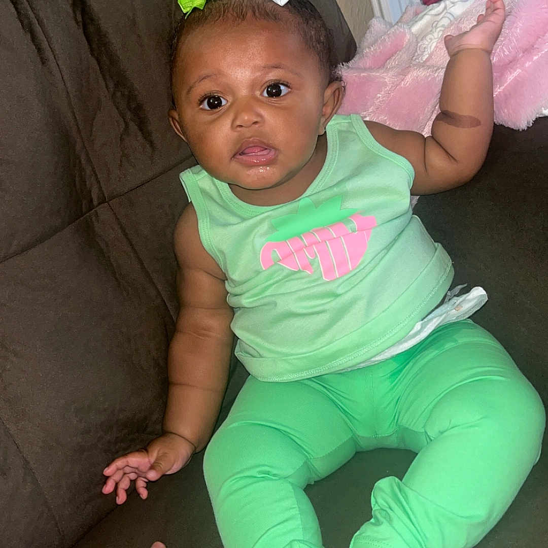 Dior is registered to the contest to win money with this photo: baby, blanket, bows, brown_couch, child, couch, curious, cute, family, green_clothing, hand, home, indoor, infant, mirror, person, pink_blanket, reflection, sitting, tattooed_hand