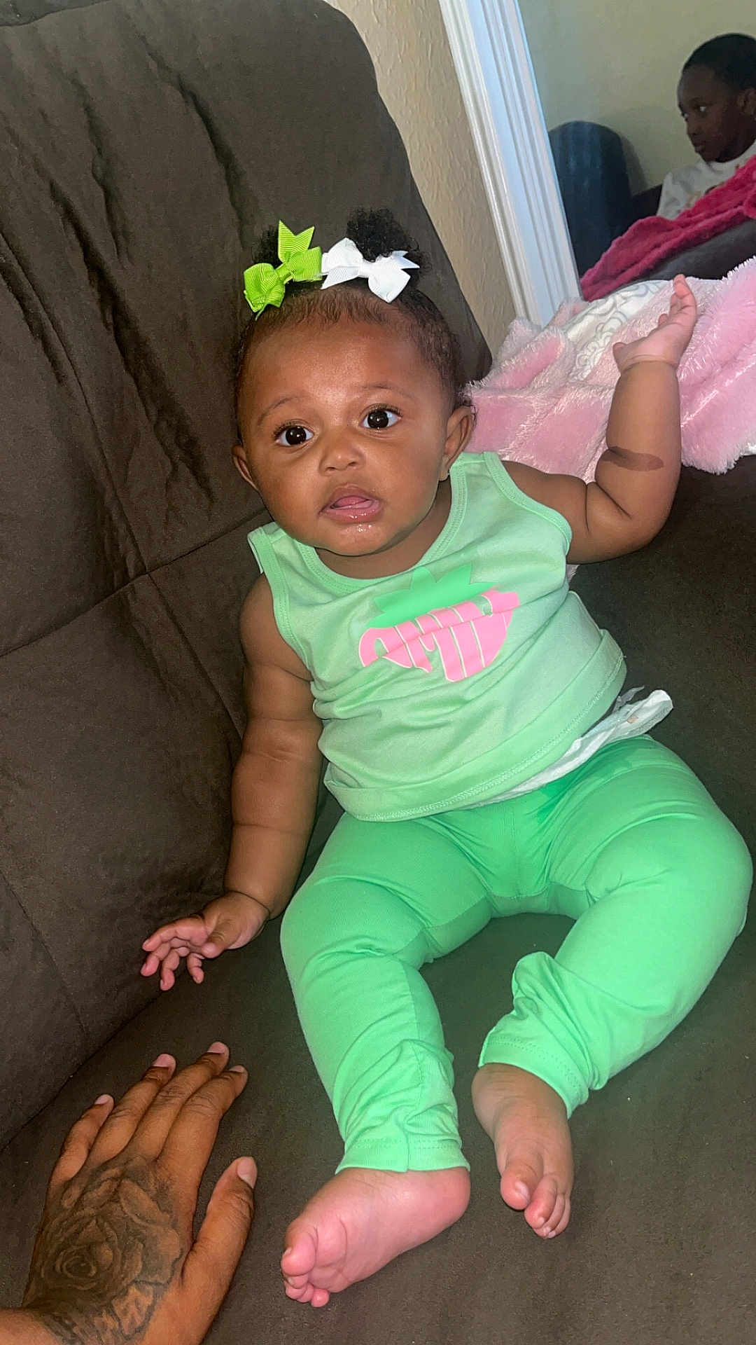 Dior is registered to the contest to win money with this photo: baby, child, infant, green_clothing, bows, couch, brown_couch, tattooed_hand, hand, reflection, mirror, blanket, pink_blanket, indoor, person, cute, sitting, curious, family, home