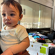 Josiah is registered to the contest to win money with this photo: toddler, child, indoor, person, white_shirt, curly_hair, holding, table, clothes, menu, blurred_background, seated, large_eyes, casual, daylight, skin, hand, face, expression, cozy
