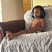 Matthew is registered to the contest to win money with this photo: child, headset, couch, blanket, living_room, indoor, person, young_child, technology, gaming, furniture, relaxing, concentration, barefoot, casual, brown_skin, short_hair, seat, comfort, home