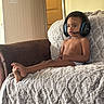child, headset, couch, blanket, living_room, indoor, person, young_child, technology, gaming, furniture, relaxing, concentration, barefoot, casual, brown_skin, short_hair, seat, comfort, home