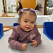 Luz is registered to the contest to win money with this photo: toddler, child, pigtails, purple_bows, sweater, smiling, face, hands, table, chair, classroom, indoor, cute, happy, person, young_child, baby, skin, hair, expression