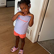 Zemirah is registered to the contest to win money with this photo: child, girl, smiling, standing, braided_hair, beads, purple_tshirt, orange_shorts, pink_slippers, wooden_floor, kitchen, oven, doorway, wall, light_switch, vent, indoor, cute, playful, home