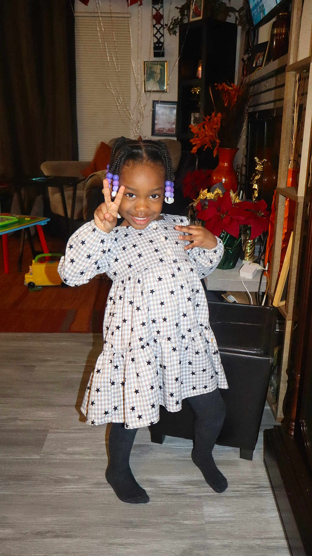 Zemirah is registered to the contest to win money with this photo: child, girl, smiling, peace_sign, dress, star_pattern, tights, indoor, living_room, floor, poinsettia, furniture, decor, hair_beads, playful, pose, happy, person, home, cozy
