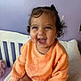 Maisie is registered to the contest to win money with this photo: toddler, child, smiling, curly_hair, peach_sweatshirt, bed, pillow, purple_wall, happy, indoor, person, cute, baby, sitting, clothing, face, portrait, young_child, expression, playful