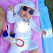 Helia participe au concours pour gagner de l'argent avec cette photo : baby, child, sunglasses, hat, towel, outdoor, plush_toy, white_clothing, pink_towel, blue_towel, grass, sunlight, cute, infant, relaxing, summer, accessory, person, leisure, toy
