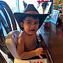 Matías is registered to the contest to win money with this photo: baby_bottle, chair, child, cowboy_hat, curtain, dining_room, expression, face, food, furniture, high_chair, indoor, mouth, photograph_frame, plant, shirtless, table, toddler, toy, window