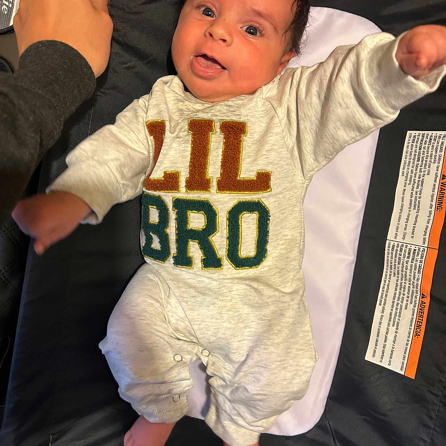 Waylon is registered to the contest to win money with this photo: apparel, baby, blanket, child, clothing, cushion, cute, face, foot, hand, happy, indoor, infant, lying_down, onesie, person, skin, smiling, text_on_clothing, young