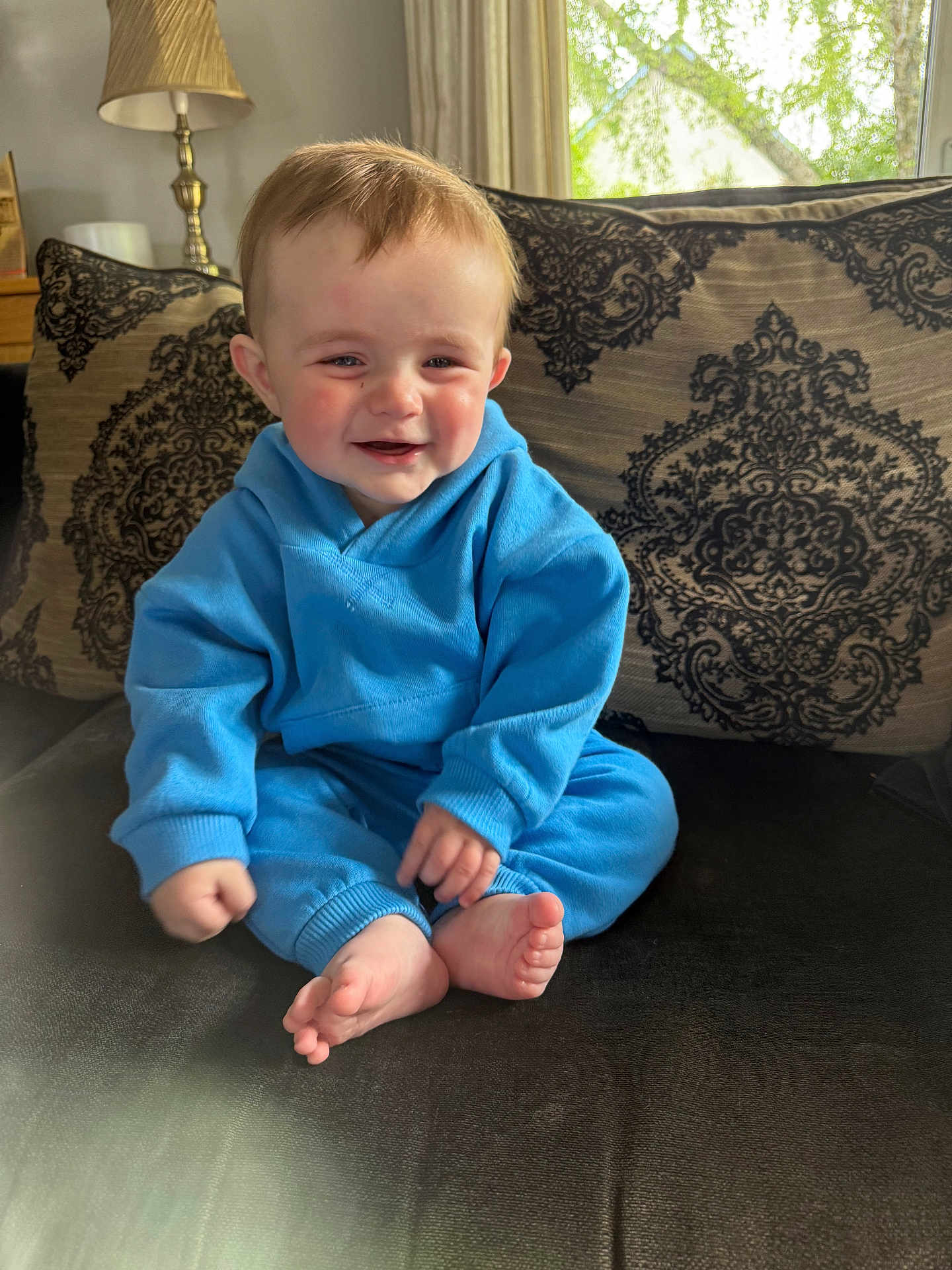 Scott joined the competition — help win amazing prizes! baby, blue_clothing, smiling, indoor, couch, cushion, patterned_cushion, child, happy, sitting, barefoot, lamp, window, curtains, person, face, home_interior, furniture, soft_light, cozy