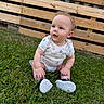 baby, child, infant, sitting, grass, outdoor, wooden_fence, backyard, onesie, smiley_pattern, shoes, white_shoes, portrait, face, blue_eyes, blond_hair, curious, happy, lawn, daylight