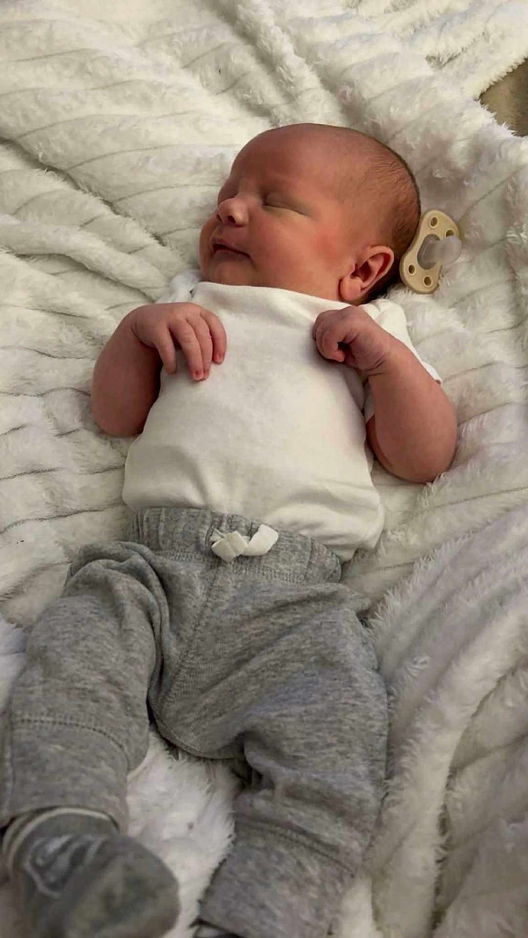 Waylon is registered to the contest to win money with this photo: baby, newborn, infant, sleeping, pacifier, onesie, blanket, soft_texture, gray_pants, sock, hands, face, head, portrait, cozy, peaceful, indoor, lying_down, closeup, adorable
