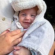 Camille a rejoint le concours — aidez-le/la à gagner de superbes lots ! baby, child, smile, hood, blanket, hand, adult_hand, knit_sweater, outdoor, nature, greenery, portrait, face, warm_clothing, cozy, infant, cute, holding, closeup, happy