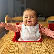 Sarah is registered to the contest to win money with this photo: baby, child, high_chair, bib, smile, happy, table, wood, indoor, person, infant, cute, pink_clothing, long_sleeve, face, cheeks, expression, sitting, arm, room