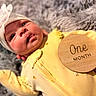 Luna is registered to the contest to win money with this photo: baby, infant, newborn, yellow_onesie, headband, bow, wooden_sign, one_month, blanket, fluffy, soft_texture, lying_down, cute, face, expression, portrait, milestone, child, clothing, indoor