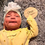 Luna is registered to the contest to win money with this photo: baby, newborn, infant, yellow_onesie, headband, flower, wooden_sign, one_month, fluffy_rug, smile, cute, lying_down, portrait, closeup, baby_clothing, soft_texture, indoor, child, person, milestone
