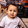 toddler, child, portrait, white_shirt, curly_hair, indoor, black_couch, wooden_floor, toy_box, playroom, smile, face, cute, young_child, eyes, sitting, floor, background_blur, happy, casual_clothing