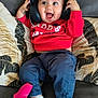 baby, headphones, red_sweatshirt, blue_pants, socks, couch, pillow, game_controller, smiling, happy, indoors, child, cute, playing, sitting, cozy, home, joyful, expression, technology