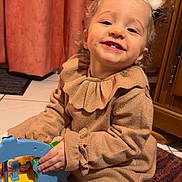 Chiara a rejoint le concours — aidez-le/la à gagner de superbes lots ! child, toddler, smiling_child, toy, headband, antler_headband, sweater, beige_clothing, curtain, indoor, playtime, hands, dimple, white_tights, carpet, tile_floor, wooden_cabinet, close_up, warm_lighting, happy_face