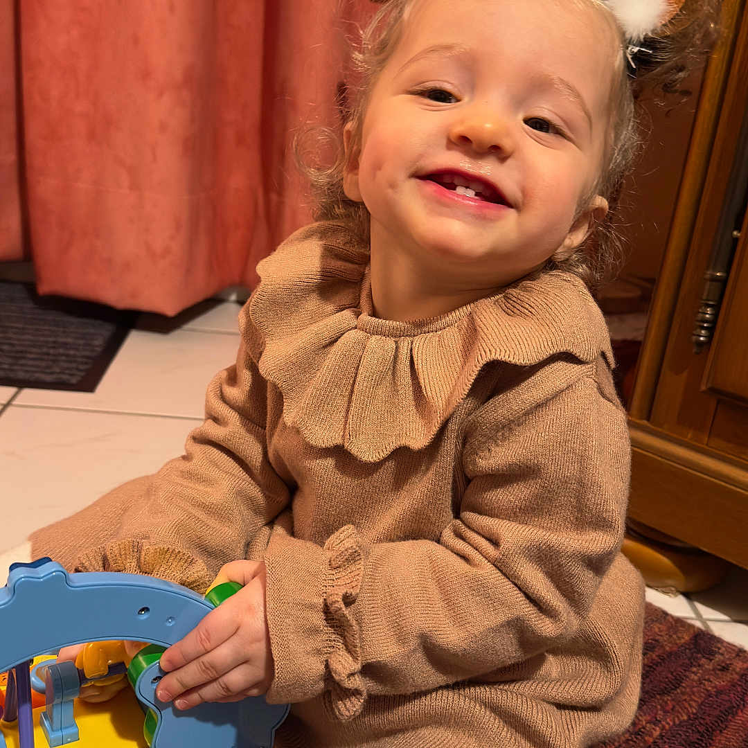 Chiara a rejoint le concours — aidez-le/la à gagner de superbes lots ! antler_headband, beige_clothing, carpet, child, close_up, curtain, dimple, hands, happy_face, headband, indoor, playtime, smiling_child, sweater, tile_floor, toddler, toy, warm_lighting, white_tights, wooden_cabinet